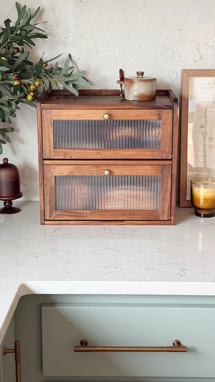 A wooden two-drawer bread box with glass panels sits on a white marble kitchen countertop.