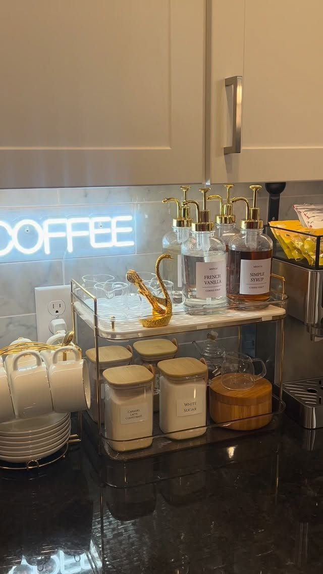 Coffee station setup with syrup dispensers, glass cups, and storage jars under a neon sign.