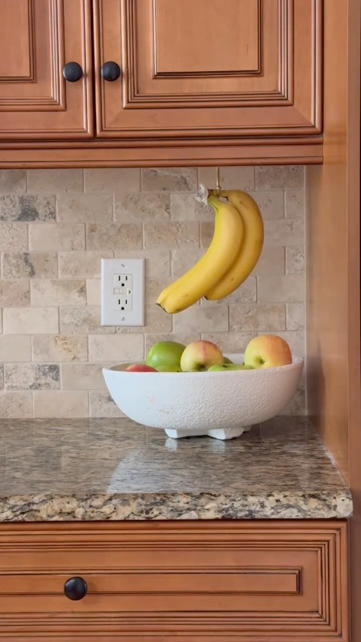 Bananas hanging from a cabinet above a white bowl of apples on a granite countertop.