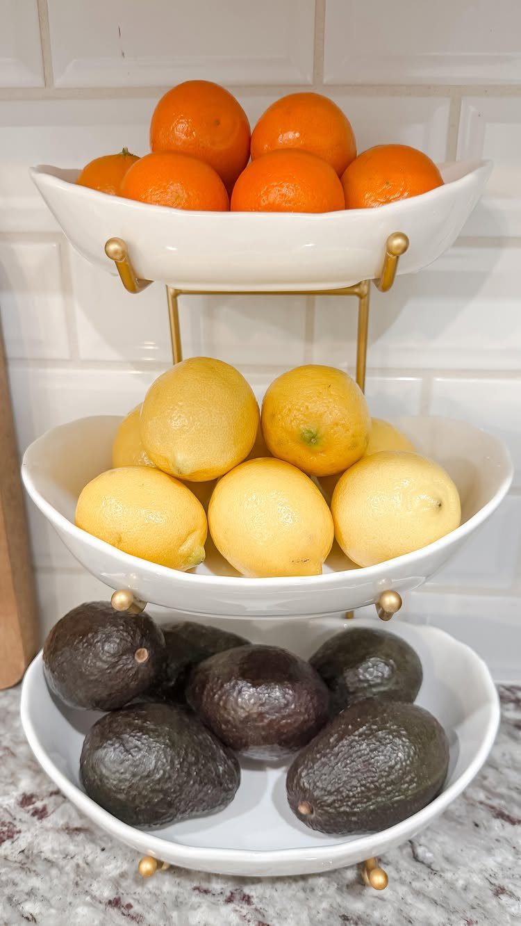 A three-tiered white ceramic stand filled with oranges, lemons, and avocados on a kitchen countertop.