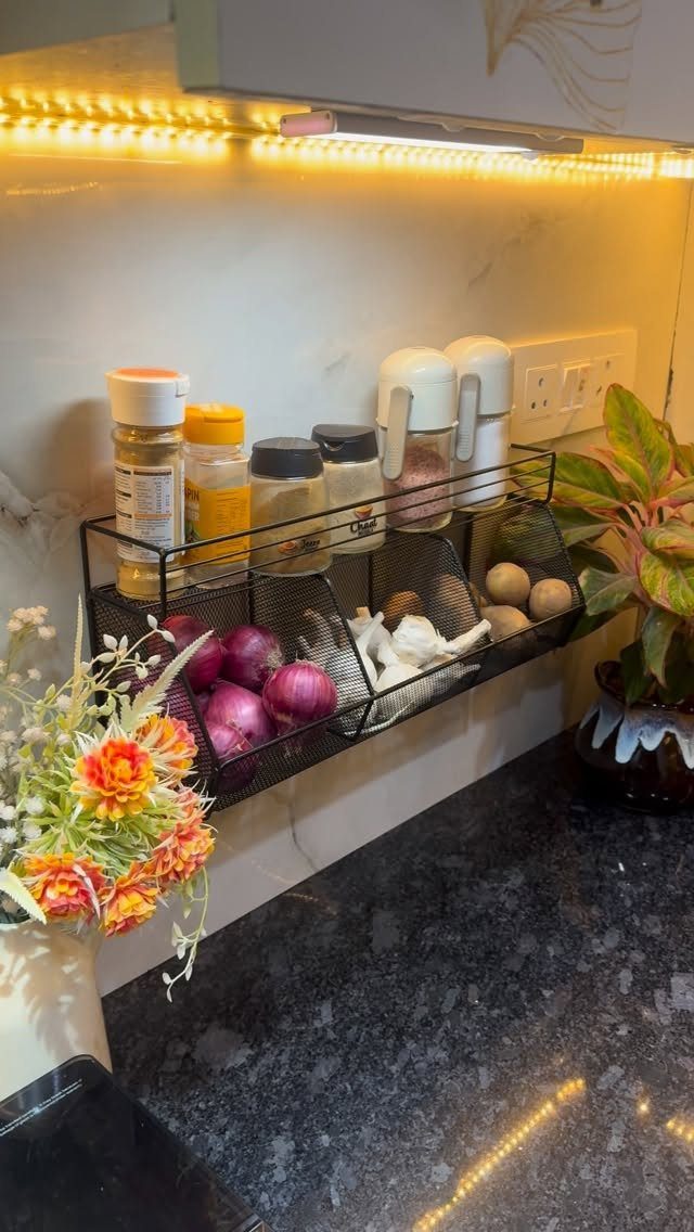 Black wire storage shelf mounted on a kitchen backsplash holding spices, garlic, and red onions.