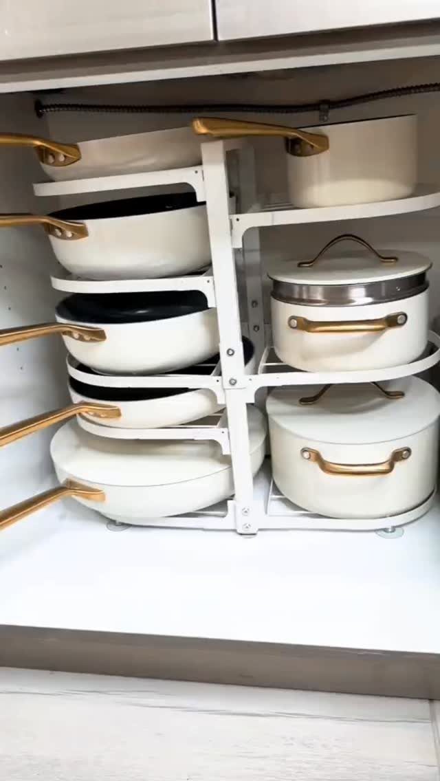 White adjustable cookware racks holding several white pots and pans with gold handles in a cabinet.