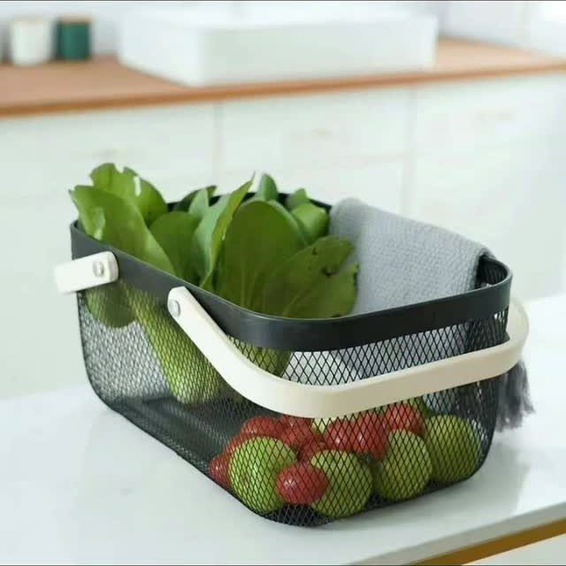 A black mesh storage basket filled with fresh green vegetables and red tomatoes on a counter.