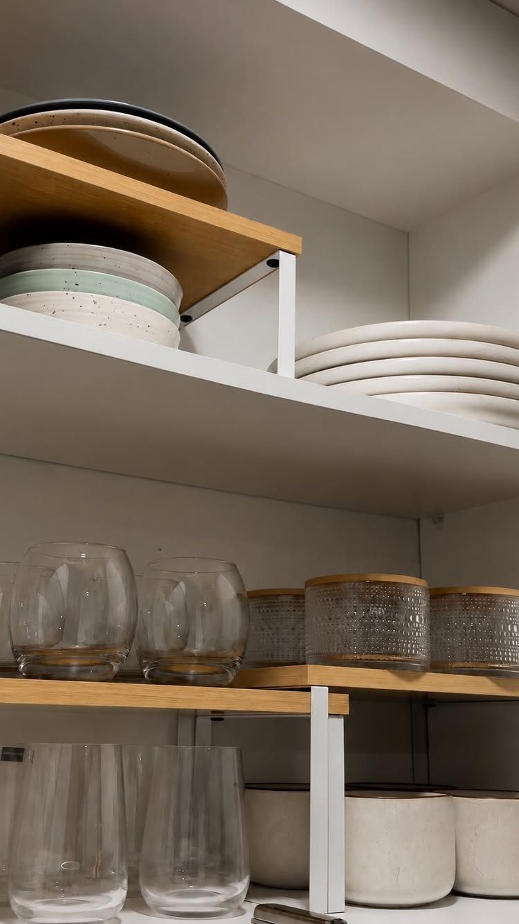 Wooden shelf risers holding stacks of plates and bowls inside white kitchen cabinets.