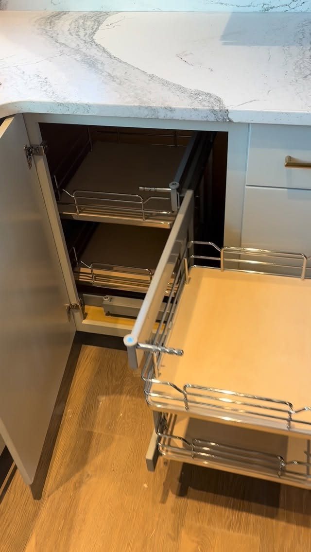 Metal pull-out wire shelving units installed inside an open kitchen cabinet under a marble countertop.