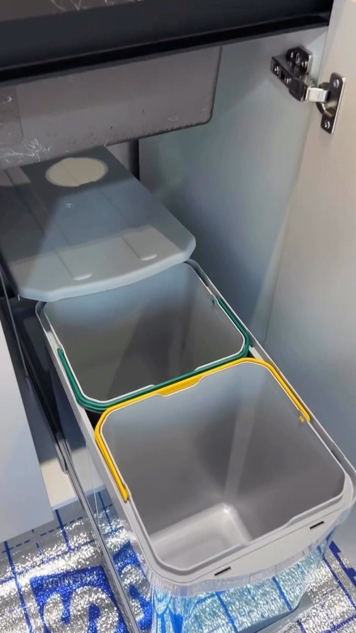 Two grey pull-out trash bins with colored rims on a sliding metal track inside a cabinet.