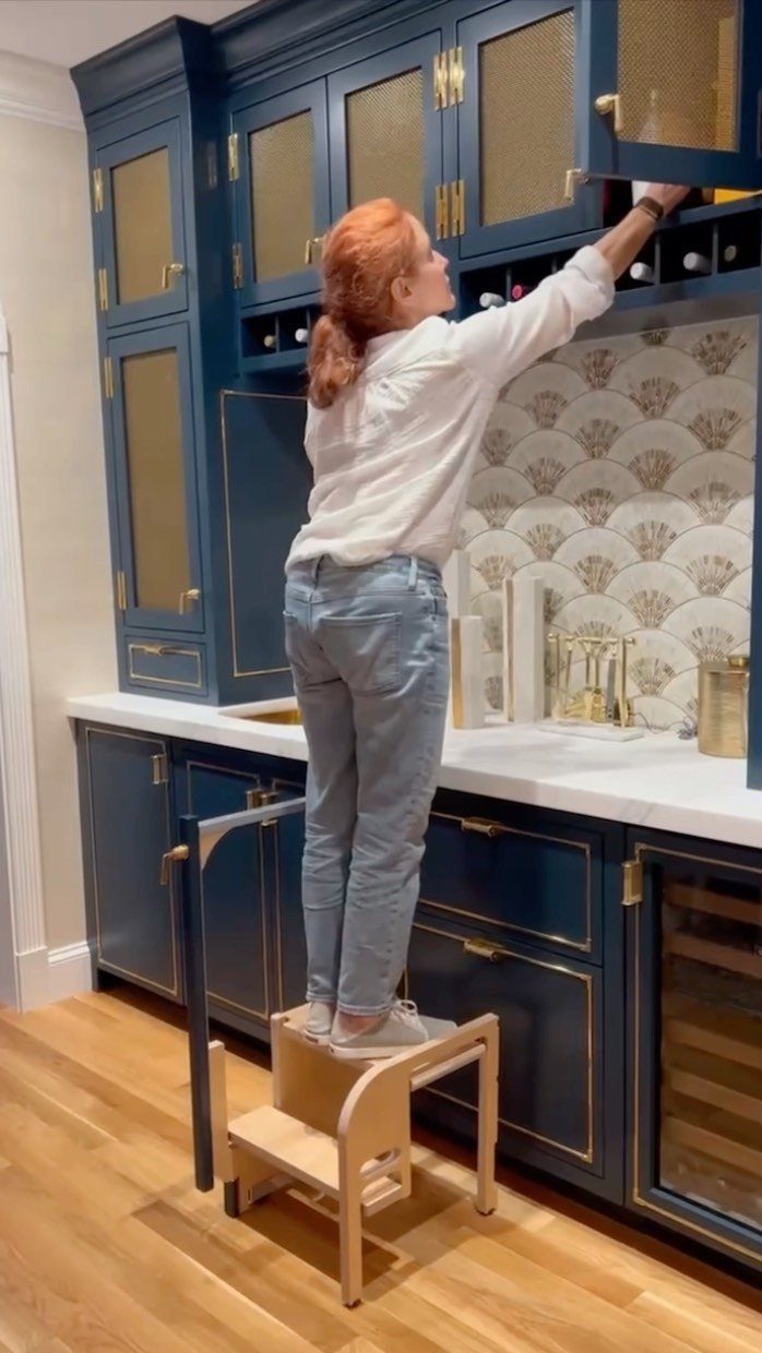 A woman standing on a wooden step stool to reach items in high kitchen cabinets.