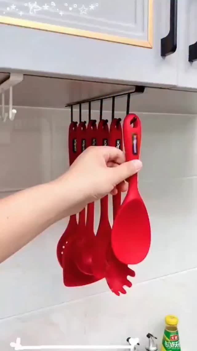 A hand grabs a red silicone spatula hanging from a rack under a kitchen cabinet.
