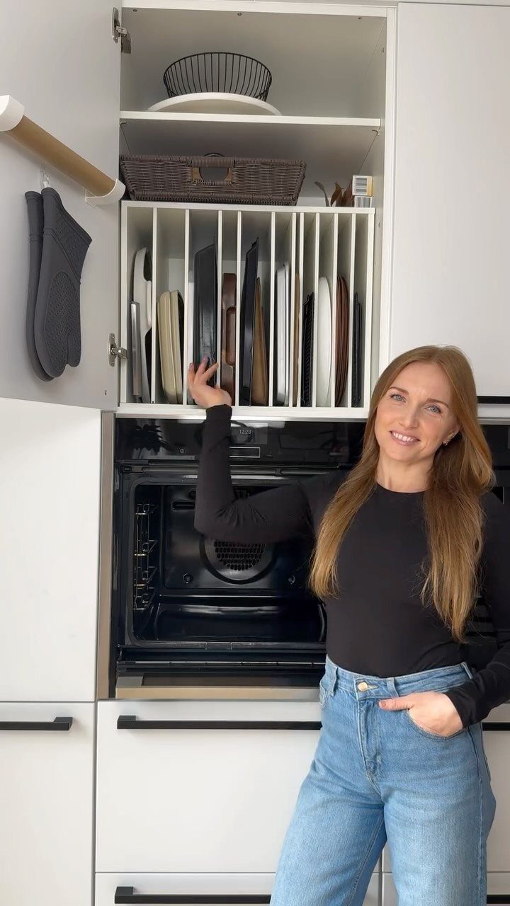 A woman stands by an open kitchen cabinet containing a vertical organizer for baking sheets.