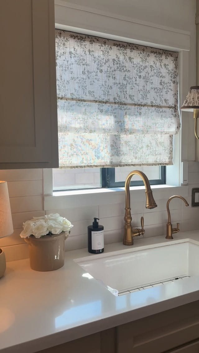 Patterned Roman shade covering a kitchen window above a white sink and brass faucet.