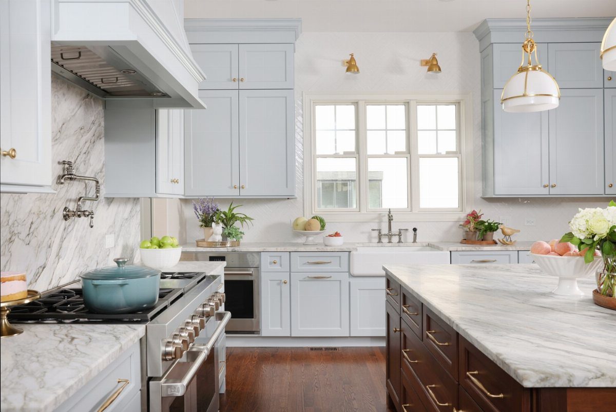 Light blue kitchen cabinets with marble countertops and brass wall sconces above a window.