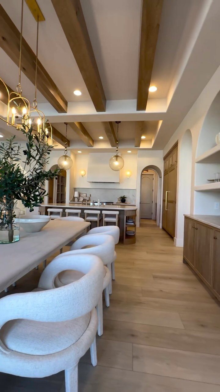 White upholstered dining chairs arranged around a long light-colored table under wooden ceiling beams.