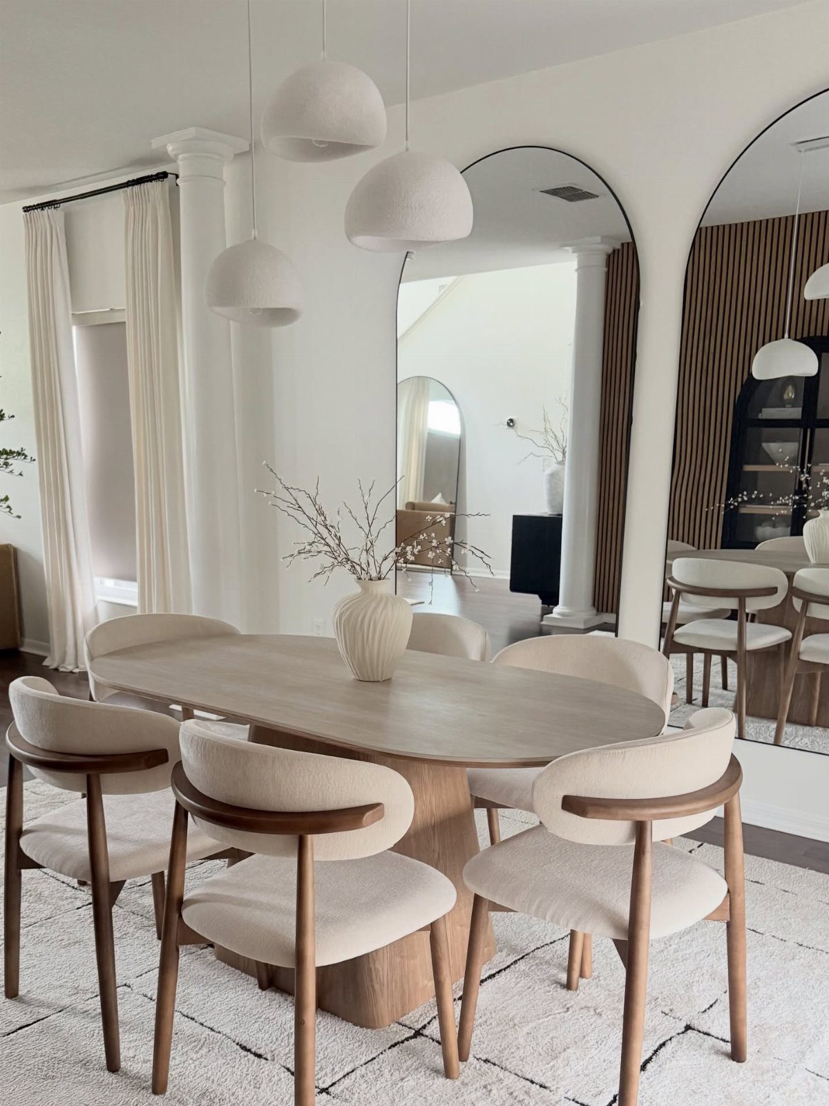 Cream upholstered dining chairs with wooden armrests surround an oval wooden table in a bright room.