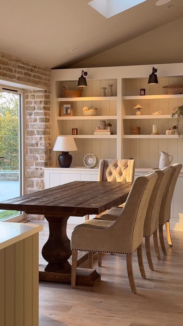 Large dark wood pedestal dining table surrounded by beige upholstered chairs in a bright room.
