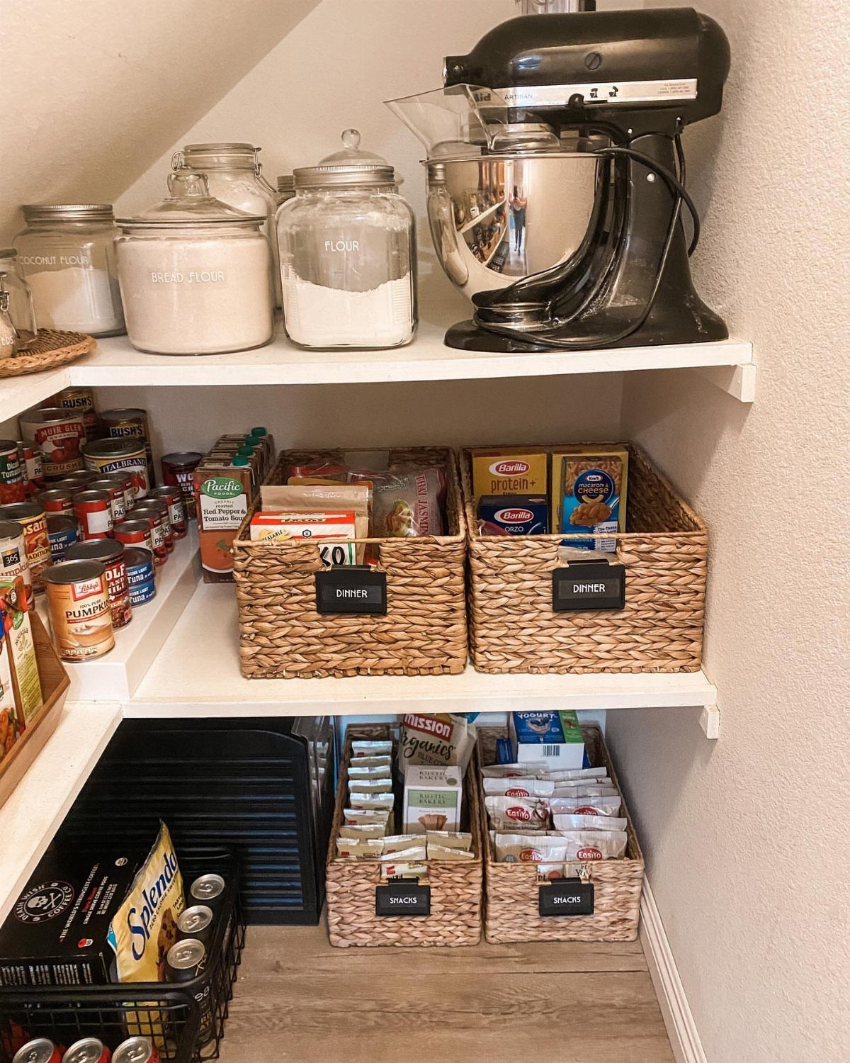 White pantry shelves under a sloped ceiling holding jars, a mixer, and woven storage baskets.
