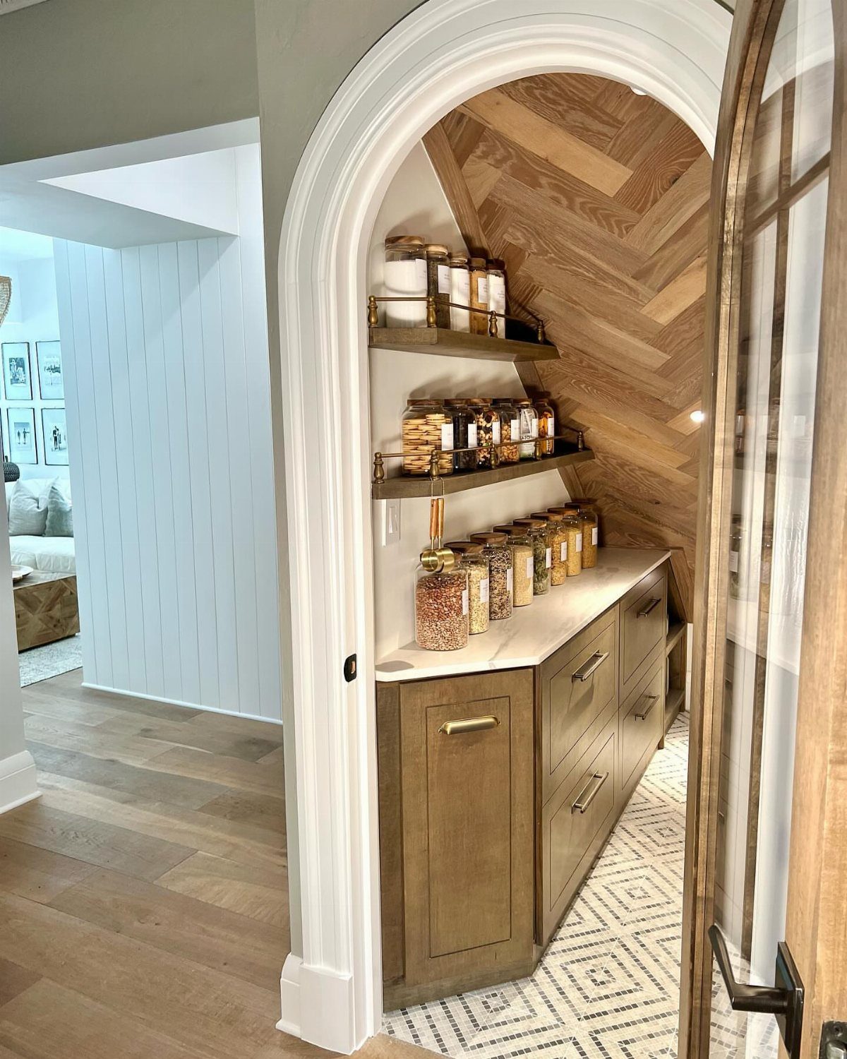 A walk-in pantry with arched entrance, wooden herringbone walls, and organized glass jars on shelves.