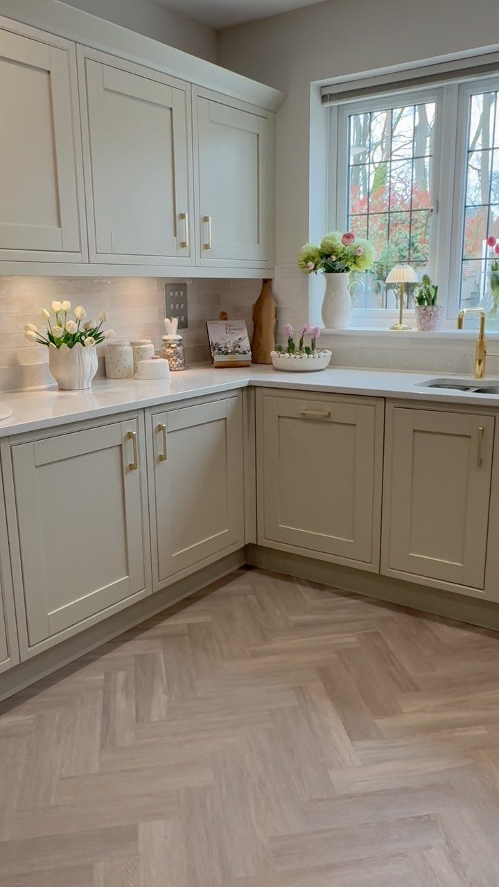 Light grey kitchen cabinets with gold handles and herringbone wood flooring near a window.
