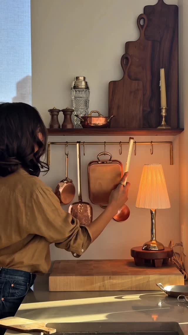 A person reaching for copper cookware hanging from a brass rail in a warm kitchen setting.
