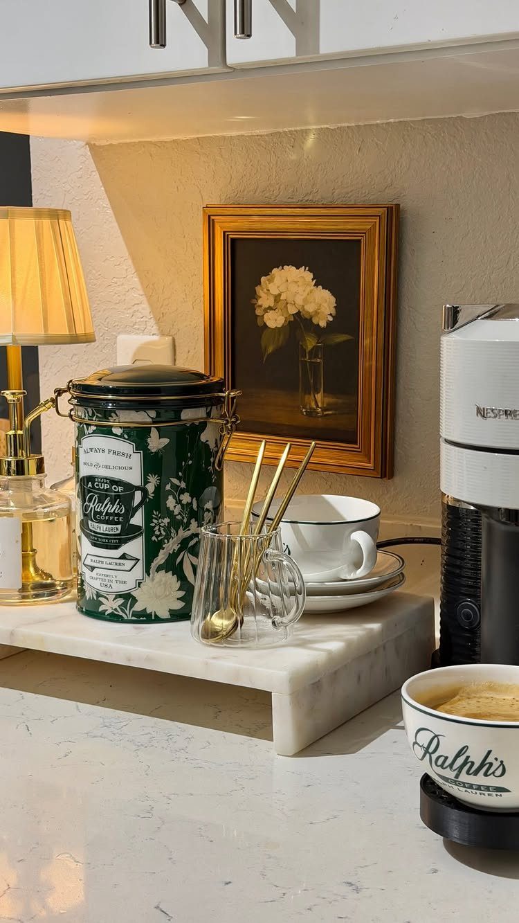 Kitchen counter decor featuring a green canister, small lamp, framed floral art, and white mugs.