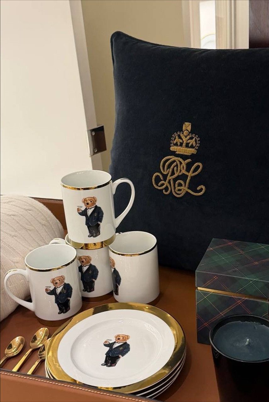 Ceramic mugs and plates with teddy bear illustrations and gold rims next to a navy pillow.