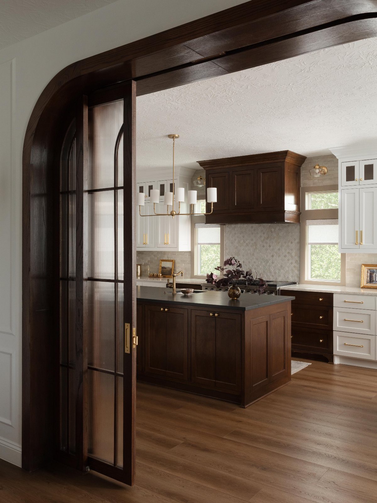 Dark wood kitchen cabinetry seen through a large arched wooden doorway with glass panes.