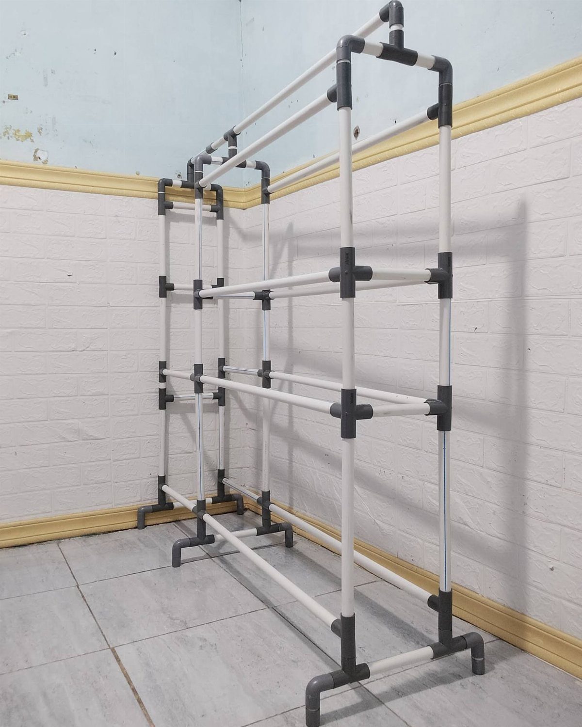 A white PVC pipe shelving unit with black joints standing in a corner against a brick wall.