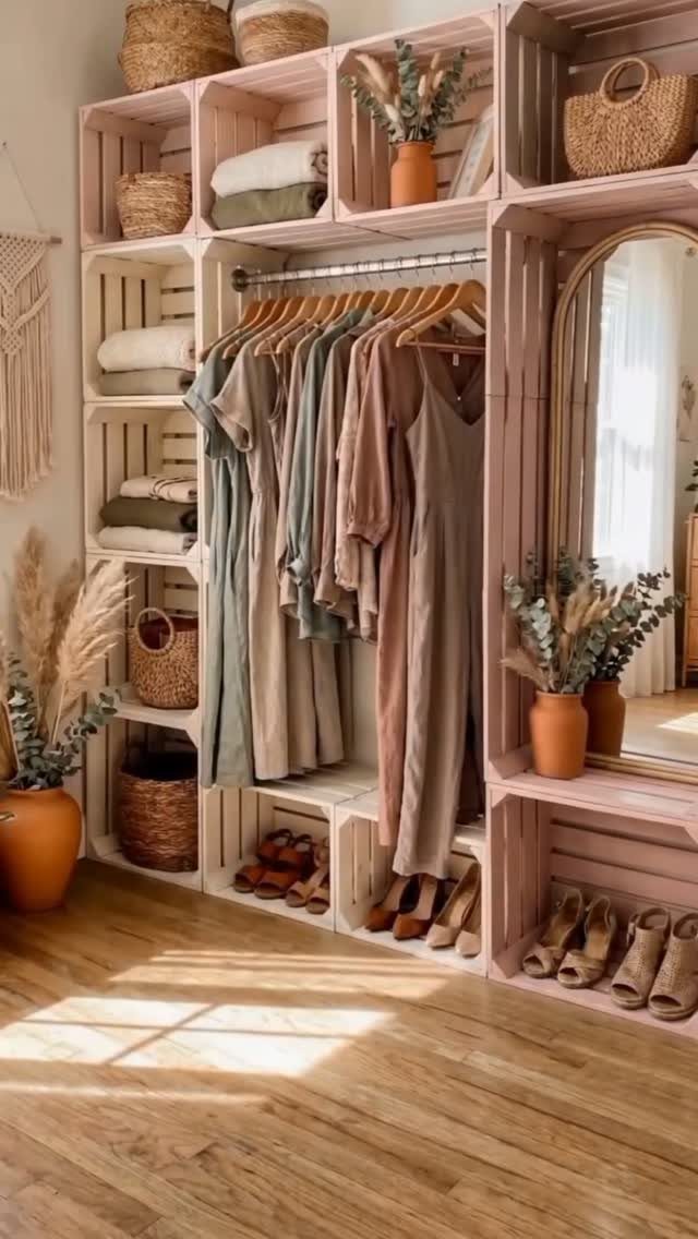 Pink wooden crate shelving unit holding clothes, baskets, and dried flowers in a bright room.
