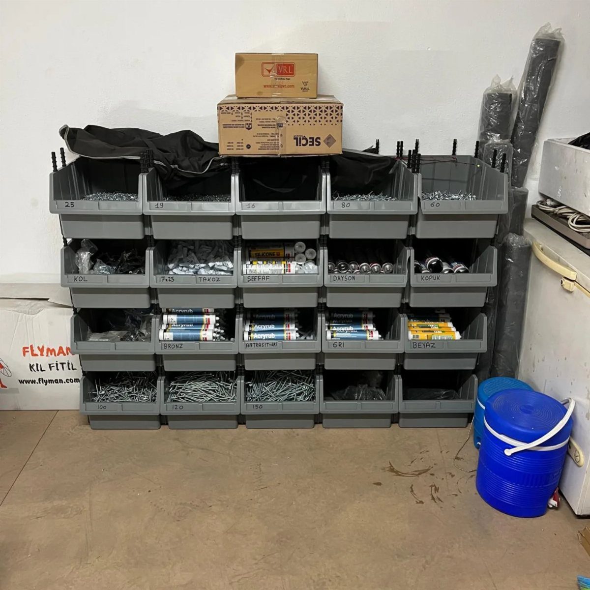 Grey plastic storage bins filled with hardware organized in a tiered shelving unit against a wall.