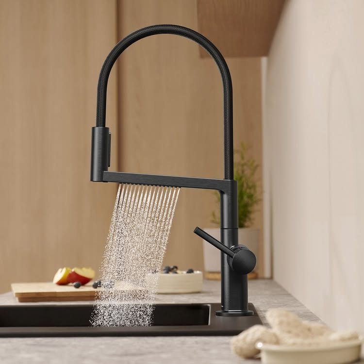 A matte black kitchen faucet spraying a wide curtain of water into a dark sink.