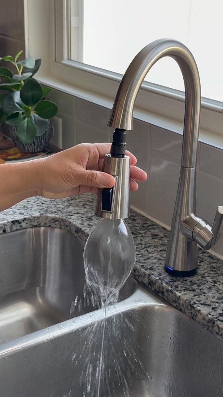 A hand operates a pull-out sprayer faucet with water flowing into a stainless steel sink.