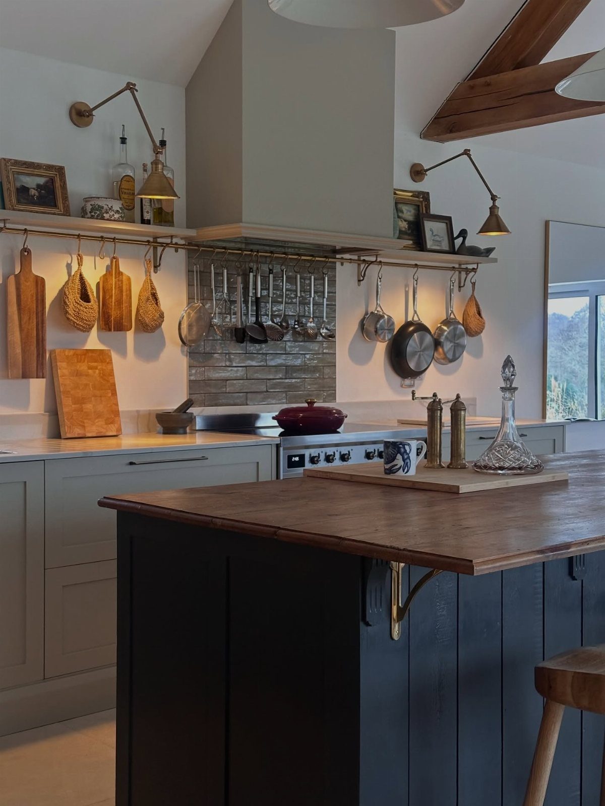 A rustic kitchen with grey cabinets, a blue island, wooden countertops, and hanging cookware.