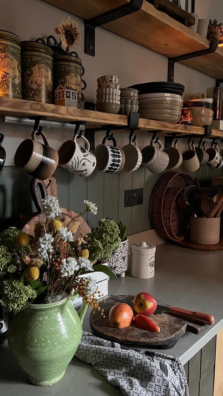 Wooden open shelves holding ceramic mugs and kitchen canisters above a countertop with flowers and fruit.